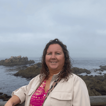 Emma is leaning against a railing with the sea behind her. She is wearing a pink top and pale pink jacket. She has curly hair.